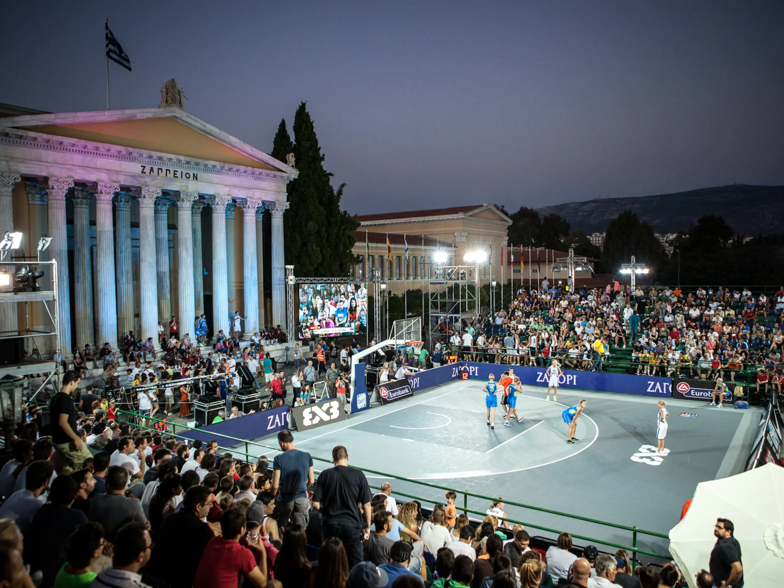 Pista de baloncesto 3x3 en una plaza urbana al anochecer, rodeada de público